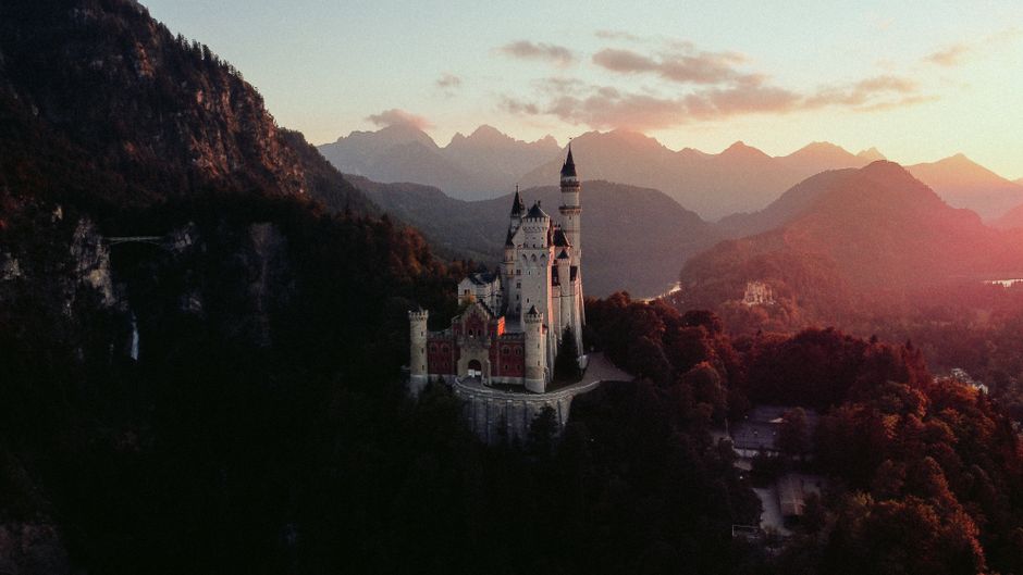 A breathtaking aerial view of Neuschwanstein Castle surrounded by stunning mountain scenery during sunset.